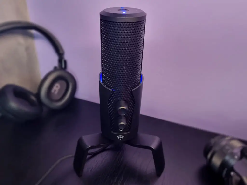 microfone GXT 258 Fyru LED USB 4 em 1 em em uma mesa preta, com fones de ouvido e câmera fotográfica visíveis, com uma parede lilás no fundo